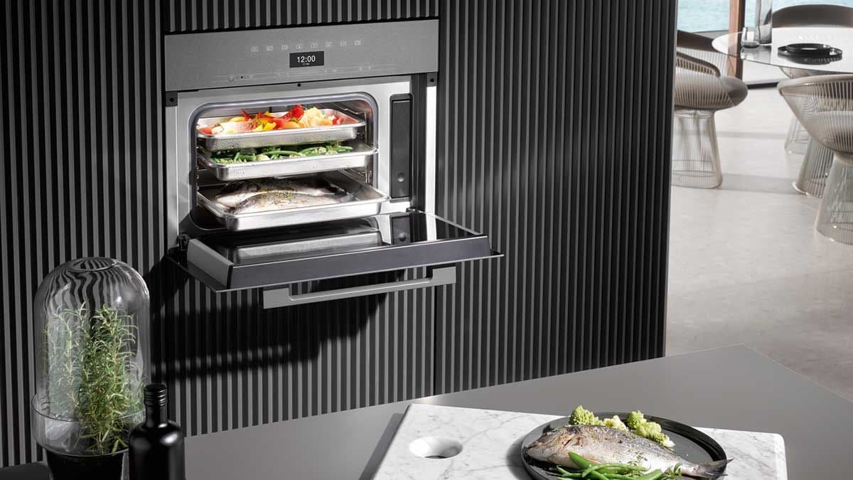 Miele Combi-Steam offen mit drei Gemüse-Schalen in der Hochschrank-Front, Indoor-Premium-Setting mit Marmor-Insel und Fisch im Vordergrund